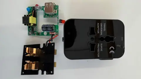 Electrical Safety First A circuit board and small plastic cover on a table beside a black, plastic travel adaptor. Text on the adaptor case, next to its sockets, reads "TRAVEL UNIVERSAL ADAPTOR" and on the other side: "SURGE PROTECTOR". A "CE" certification standard symbol is also displayed on the casing.
Copyright