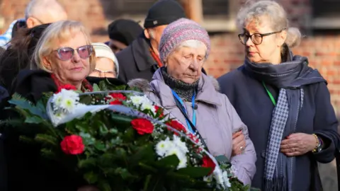 Reuters Three women attend a wreath-laying ceremony to mark 80 years since the liberation of Auschwitz; one holds flowers, another looks down and the third looks off to the side.