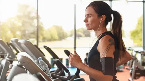 A woman uses a treadmill in a leisure centre. She is listening to music and has headphones in her ears. There is a large window behind her with open space and trees and grass.