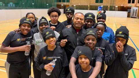 Gaby Kitoko, who is the centre of the picture and wearing a blue jacket with a silver zip, stands with just some of the youngsters who form part of the group. Most of them are wearing blue shirts that say 'Chance to Shine/Street' on them, and they are standing on a pitch they have just completed a game on.