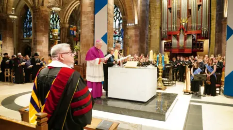 PA Media Clergy members lead a ceremonial service at the altar inside St Giles' Cathedral in Edinburgh during the Royal Kirking of Parliament. Choir members and musicians stand near a large pipe organ in the background, while representatives from different faiths and civic groups hold programmes along the side aisle. A man in ceremonial academic robes sits in the foreground beneath the cathedral’s stone arches and stained glass windows.