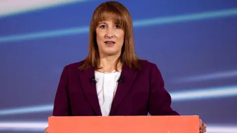 PA Media Rachel is standing in front of a salmon-pink lectern, wearing a dark maroon blazer and white shirt with straight brown shoulder-length hair.