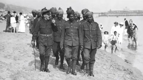 Getty Images In a black and white photo from 1915, a dozen or so Indian soldiers walk on Bournemouth beach, accompanied by a British officer. The pier is in the background and people are paddling in the sea.