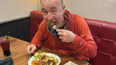 Chris Wright Runner Chris Wright is sitting at a pub table with red leather seating. He is eating a Sunday roast lunch and has a pint of squash to the right of him.