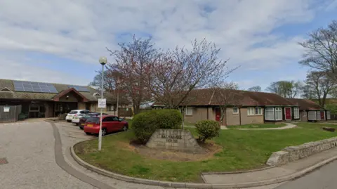 Balnagask House with its low brick buildings, parked cars and grassy grounds. A stone sign surrounded by shrubs reads “Balnagask House.”