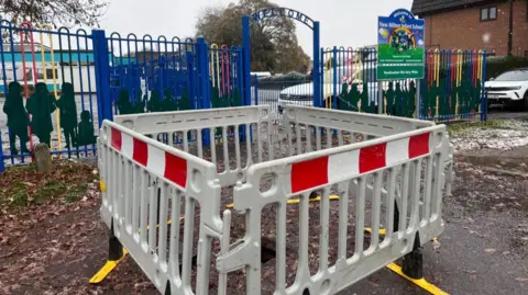 Plastic fencing surrounds a small hole in the ground outside the entrance of New Milton Infant School