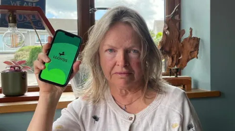 Annabel Yates from Crackington Haven in Cornwall is sat down looking straight at the camera. She has a frown on her face and is holding up a phone with the Lloyds loading screen on it. 