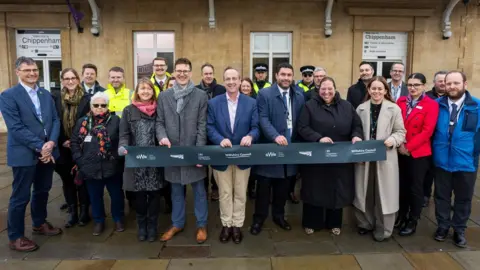 A group of people stood on a pavement smiling widely with a train station behind them