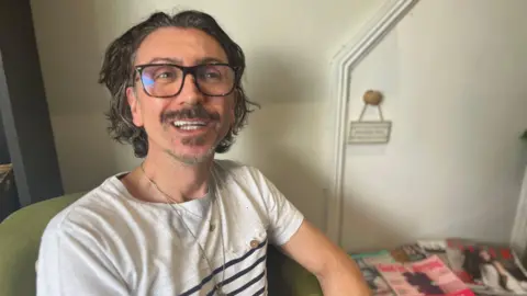 Julia Gregory/BBC Dennis Macco is in his hair salon wearing a white T-shirt with black stripes and a chain around his neck. His hair is pushed back and he has glasses. He is looking up and smiling at the camera. There are magazines on a coffee table to one side of him and there is a cupboard with a sign on the handle.