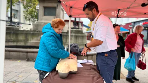 A man is teaching a woman how to give CPR. The woman has red hair and is wearing a blue coat. She is stood in front of a table and is placing both of her hands on the chest of a CPR dummy. The man is demonstrating with his hands how to perform CPR.