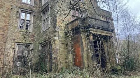 Photograph of the entrance to the derelict Winstanley Hall in Wigan. The images shows overgrown trees and  broken windows.