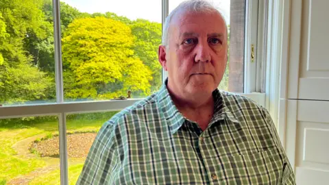Jonny Humphries/BBC Alan Jones, 59, who has thinning grey hair and is wearing a green chequered short-sleeved shirt, sits by a first floor window looking out onto a green garden lined with trees. 