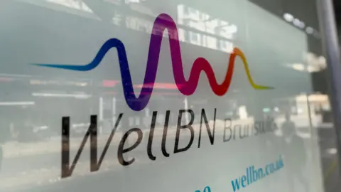 An image of the WellBN clinic sign in Brunswick, Brighton. In the window you can see a reflection of a child walking passed and some shops on the road opposite.
