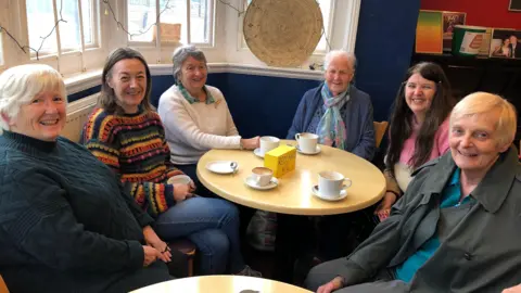 A group of six women are sat around a round coffee table with cups of coffee in front of them.  There are smiling at the camera.  Also on the table is a yellow block which reads Redlands WI.