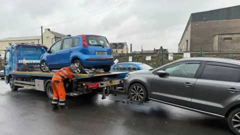 Bradford Council A truck with a blue car on it and towiung a grey car with a council worker in high viz clothing