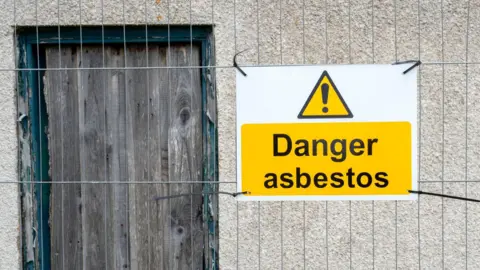 A yellow sign saying danger asbestos, tied to a metal fence