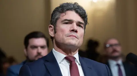 Kent during a committee hearing, wearing a dark blue suit and a red tie, with a serious expression