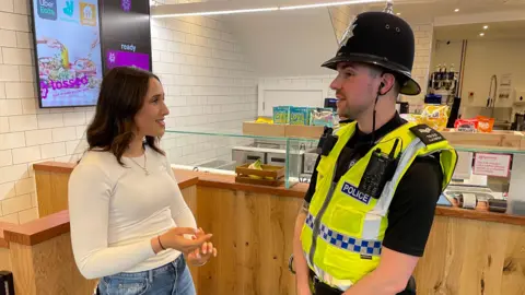 Tara is speaking to Sergeant Kemish in front of the food counter 
