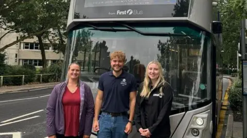 Two women and a man standing in front of a grey Norwich electric First Bus