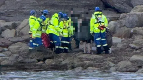 HM Coastguard Six coastguards, wearing yellow hi-vis jackets and blue trousers, is assisting several people, whose faces are pixelated. They are standing beneath a cliff. 