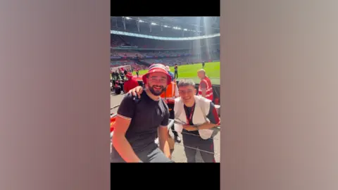Adam Paterson One man is looking at the camera, wearing a red bucket hat and black t-shirt. Another man is bending towards him, with his arm around his shoulder. Both are posing as one sits, and the other stands in a football stadium. A football pitch can be seen behind them. 