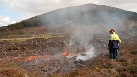 Two men standing in the Scottish countryside as a fire burns in the brown grass