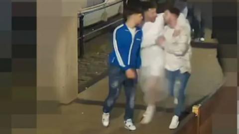 A CCTV image of three men walking under a covered section of Bristol's Harbourside. They all have their arms around each other and are laughing. The man in the middle appears to be wearing a white wedding dress, while the two men either side are in jeans and casual clothes  
