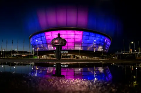 The OVO Hydro arena in Glasgow is lit in purple and blue at night, reflected in a puddle in the foreground, with a large sculpture standing outside the building.