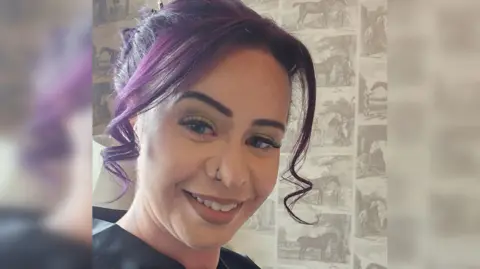 Tattoo Morningstar A woman with purple hair smiling at the camera she has her nose pierced and wears a black top.