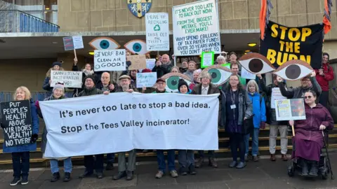 About two-dozen protesters standing on the outside steps of a council building with blue and yellow badge. The protesters, of all different ages, are holding placards and signs. One sign says "Stop the burn" in yellow lettering on a black flag. The largest banner at the front of the group says "It's never too late to stop a bad idea. Stop the Tees Valley incinerator". The banner is white. Lots of people are holding pictures of eyes. 