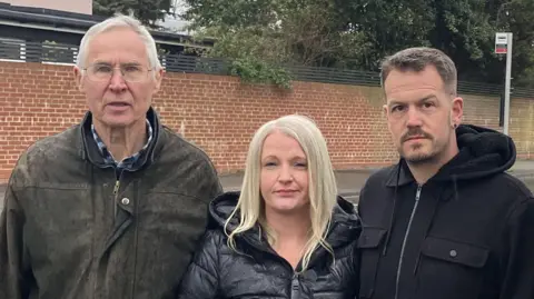 Two men and one woman standing outside. They do not look impressed and are all looking at the camera. There is a road, a brick wall and a bus stop behind them.