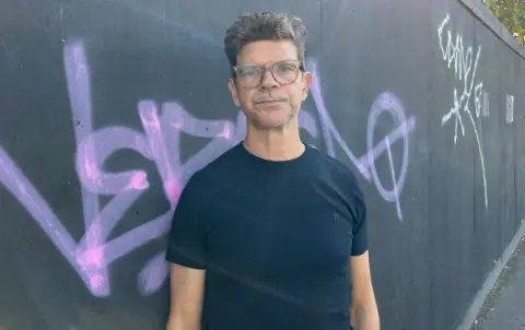 A man with grey-black hair looks at the camera. He is bespectacled and is wearing a blue T-shirt