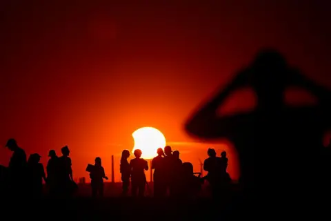Ina Fassbender/AFP/Getty Images An orange sky with the Sun in the centre, there is a small bite sized chunk taken out of its left side as the Moon covers the surface. People standing around in groups with telescopes are in shadow