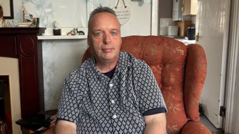 BBC A man in an orange chair has dark hair swept back. He is wearing a black and white shirt
