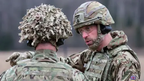 PA Media A man in military uniform, goggles and a helmet points while in conversation with troops.