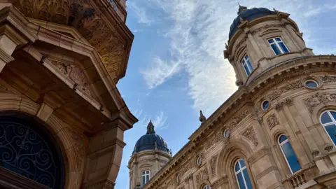 Isle Explorer Two stone buildings pictured against a sunny sky with wispy clouds. The buildings are sandstone and date from the Victorian era. They have a wealth of detail, including arched and circular windows and stone balustrades