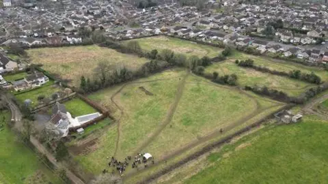 People For Packsaddle An aerial view of a several green fields with paths crossing them and hedges along the borders. A small crowd of people are standing in one corner of a field. In the distance, many houses can be seen, on the edge of Frome.