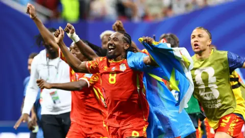 Reuters A group of DR Congo players celebrate and run to their fans after the final whistle against Uzbekistan. They're shirts are red and one is holding the DR Congo flag in his arms and cheering.