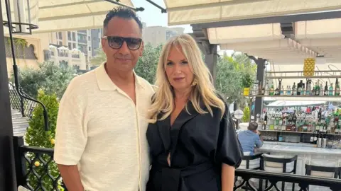 Dale Gordon Dale Gordon has black hair and is wearing black sunglasses and a cream short-sleeved shirt. He has his arm around his partner, who has long blonde hair and is wearing gold earrings and a black satin dress with lapels. They are standing under cream sun shades and there is a well-stocked outdoor bar behind them.