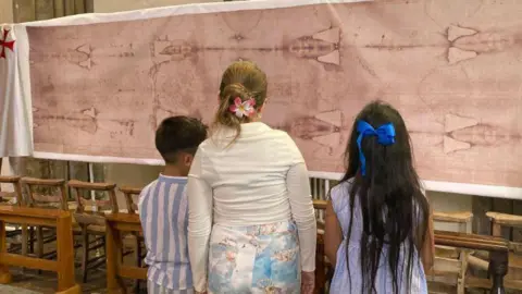 Sharon Edwards/BBC Three children stand in front of the large cloth with a faint image of a man’s body imprinted on it. The children wear light-coloured clothes and two girls have flowers or bows in their hair.
