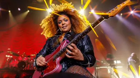 Getty Images Lizzo on stage in a black leahter jacket and fishnet tights, on her knees playing a red sparkly guitar