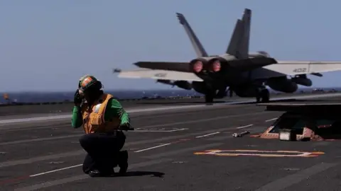 Reuters A man kneels down on the deck of aircraft carrier USS Gerald R Ford as a fighter plane is seen behind him (09/03/26)