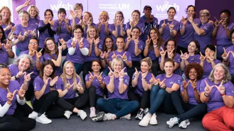 University of Hertfordshire Graduates smiling wearing purple T-shirts and making a "w" shape with their fingers