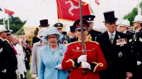 Manx National Heritage Queen Elizabeth II in a blue suit and hat with white gloves - standing behind someone in a red suit with white gloves holding a sword 