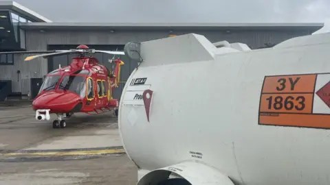 A white jet fuel tank is in the foreground with orange and black lettering, behind is the red and yellow Cornwall Air Ambulance parked outside the hangar