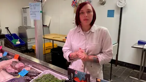 Fishmonger Steph Turley standing in front of her fish counter, wearing a white overcoat rolled up at the sleeves. She has shoulder length, straight purple hair and two lip piercings.