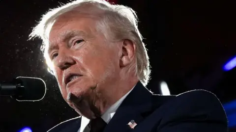 President Donald Trump is seen up close behind a podium with a mic wearing a suit and tie. The suit has a small American flag pin. He is indoors with darker lighting and he is lit from behind. He is grimacing. 