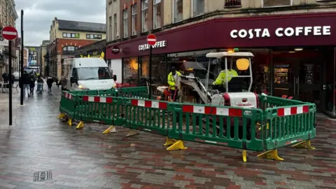 Alex Pope/BBC National Grid workers in the centre of a town, with a van and digger. There is green plastic fencing around their worksite on a pedestrianised area of a shopping street. They are next to a Costa Coffee and two "no entry" signs. 