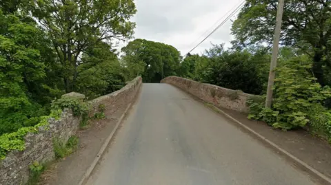 Google A Google street view image of a rural road going over a stone bridge. There is a telegraph pole on the right with wires stretching into the distance. Many trees surround the bridge and the walls of the bridge are covered in greenery. 