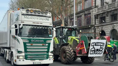 A lorry and a tractor are parked in the middle of O'Connell Street in Dublin. A sign on the front of the tractor says: "No farms, no food". The tractor is lime green in colour while the lorry is white with green detailing.
